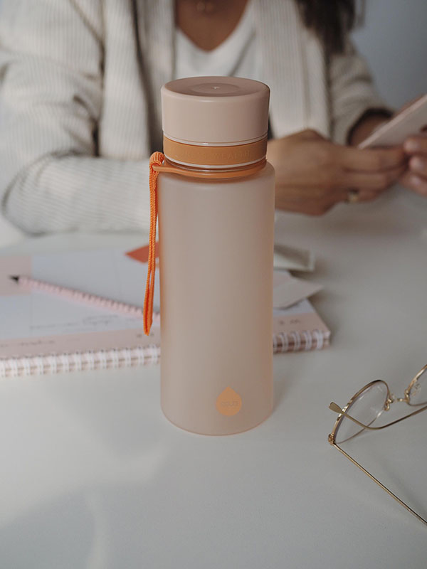 EQUA BPA FREE water bottle, Sunrise, water bottle on the office table, woman is working in the background, minimalistic design, no motif, peach color