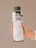 Botella de agua de cristal con tapa y soporte metálicos negros y cubierta de mármol blanco y negro para proteger la botella de agua de cristal sostenida con las manos.
