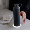 Botella de agua inteligente en color gris oscuro con un recordatorio luminoso en la parte inferior para mantenerse hidratado y una mano para coger la botella y beber el agua