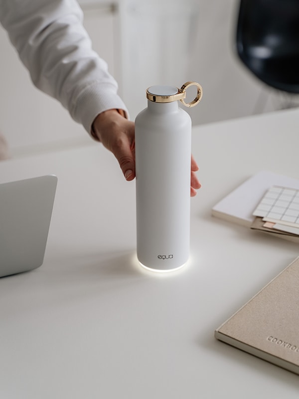 EQUA Smart Water Bottle Snow White made from Stainless Steel in White Colour and Gold Lid and Ring detail in the center of the image with hand reaching out to take it to make a sip. 