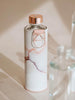 Bottiglia d'acqua in vetro lavico con dettagli in oro rosa del coperchio con logo a goccia e supporto in metallo oro rosa. La bottiglia d'acqua è protetta da un finto coperchio con design in marmo rosa.