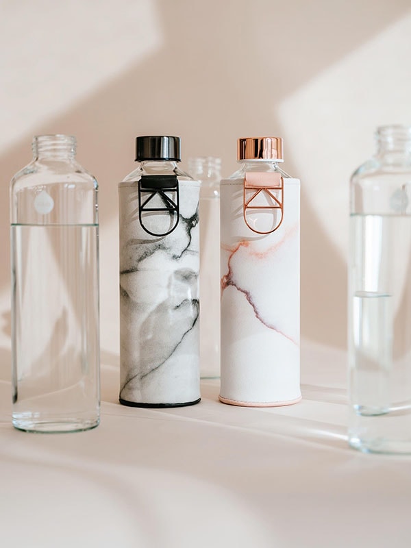 Lava Glass Water bottle with Rose Gold metal holder and lid. Glass water bottle is protected with a faux leather cover with pink marble print on off white surface.
